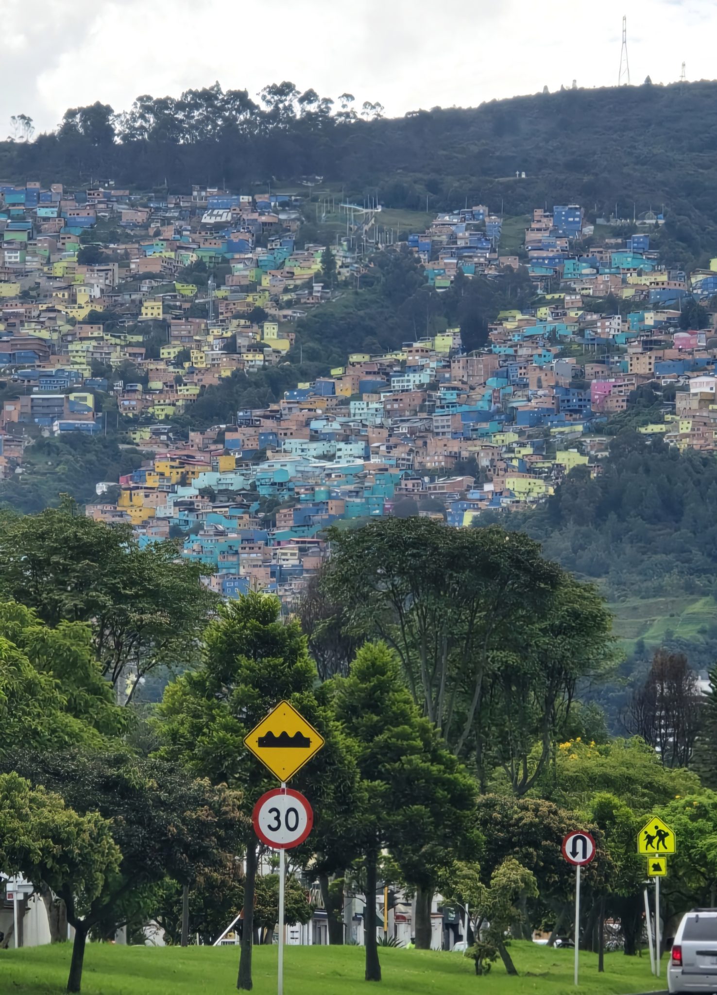 A hillside densely populated with colorful houses in various shades of blue, green, yellow, and pink. In the foreground, there are several trees and street signs, including one warning of a bump in the road and another indicating a speed limit of 30. The scenery suggests a lively urban neighborhood set against a backdrop of mountains and a cloudy sky.