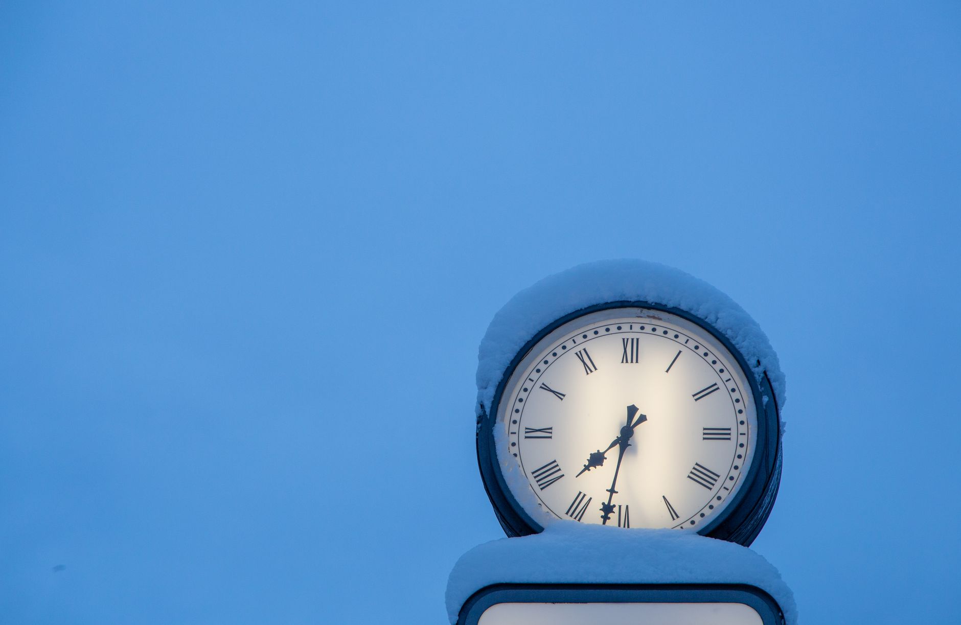 A vintage clock dusted with snow against a clear winter sky, showing Roman numerals.